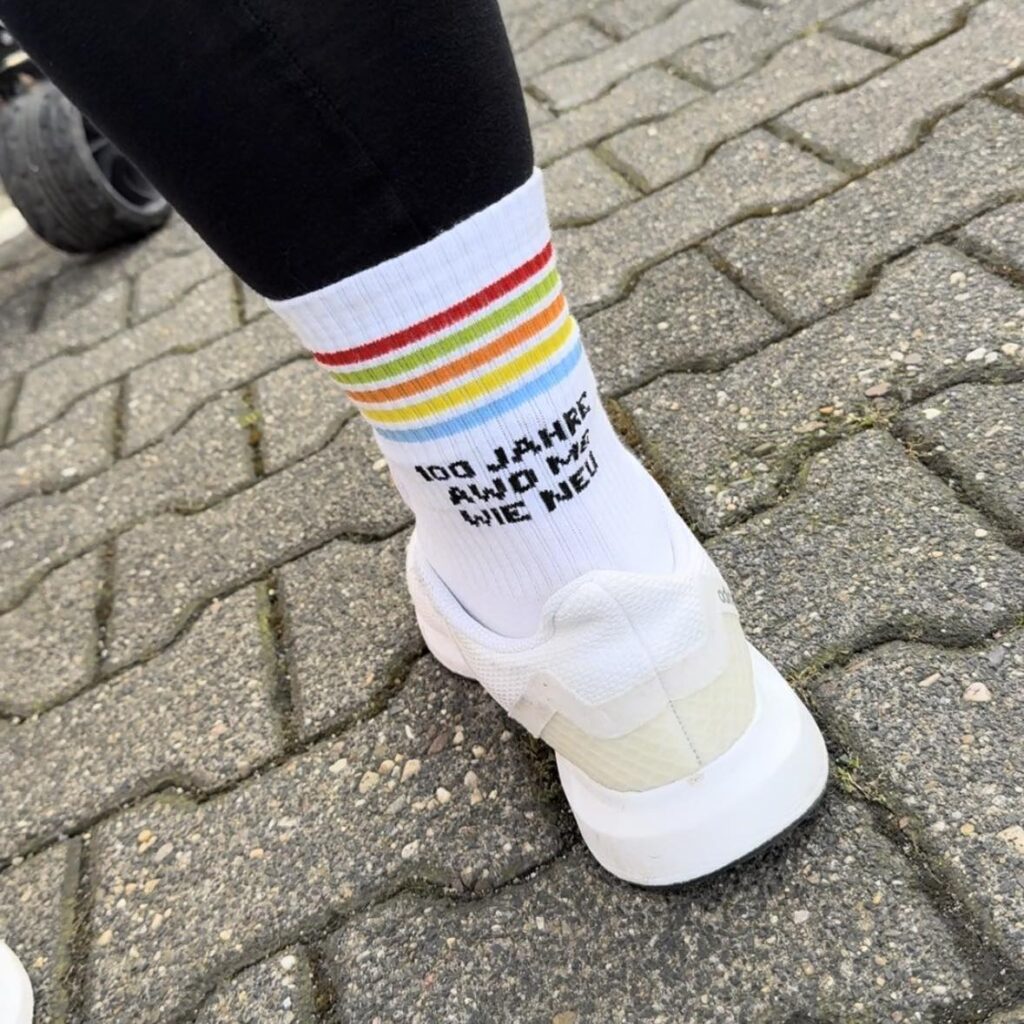 QueerPflege-LSBTIQ-Pflege Nahaufnahme einer Person, die weiße Turnschuhe und weiße Socken mit bunten Streifen und dem Text „100 JAHRE UND KEIN BISSCHEN WEISE“ auf einem Kopfsteinpflasterweg trägt.