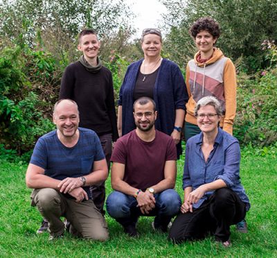 QueerPflege-LSBTIQ-Pflege Eine Gruppe von sechs Personen, drei stehend und drei kniend, posiert draußen auf einer Rasenfläche mit Bäumen und Büschen im Hintergrund für ein Foto.