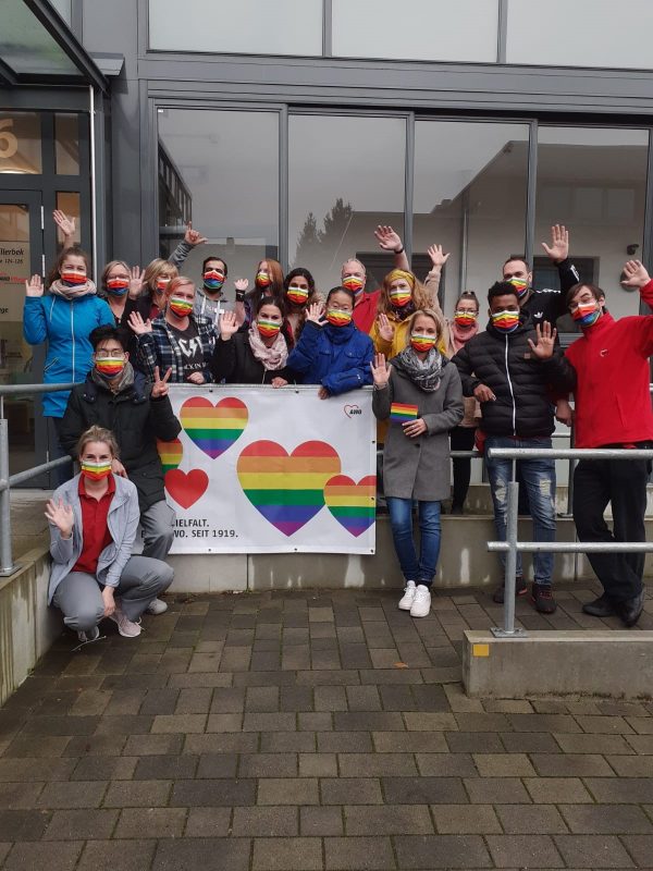 QueerPflege-LSBTIQ-Pflege Eine Gruppe von Menschen mit Regenbogen-Gesichtsmasken steht vor einem Gebäude, hält ein Banner mit regenbogenfarbenen Herzen und winkt in die Kamera.