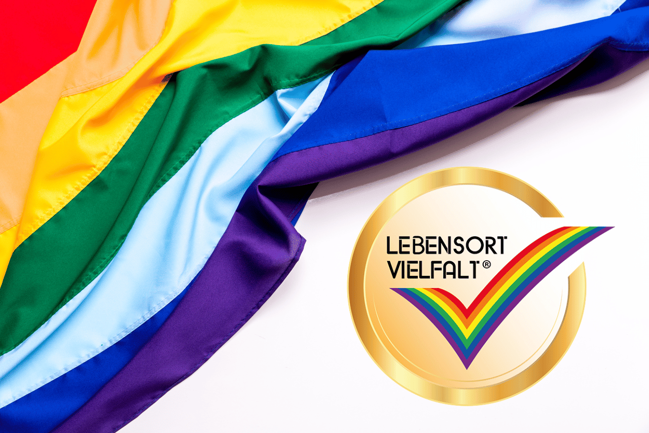 QueerPflege-LSBTIQ-Pflege Ein bunter Regenbogenstoff und ein goldener Button mit einem Regenbogenhäkchen und dem Text „Lebensort Vielfalt®“ symbolisieren die queerfreundliche Pflege.