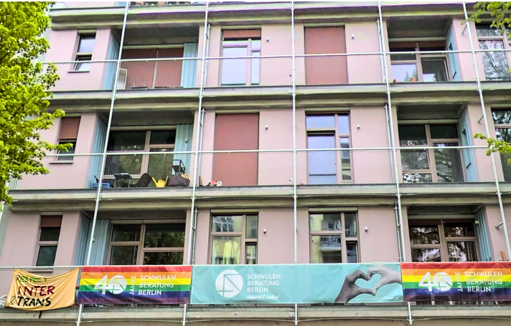 QueerPflege-LSBTIQ-Pflege An den Balkongeländern eines mehrstöckigen Gebäudes hängen Banner in Regenbogenfarben und mit Aufschriften wie „40 Schwulen Beratung Berlin“.