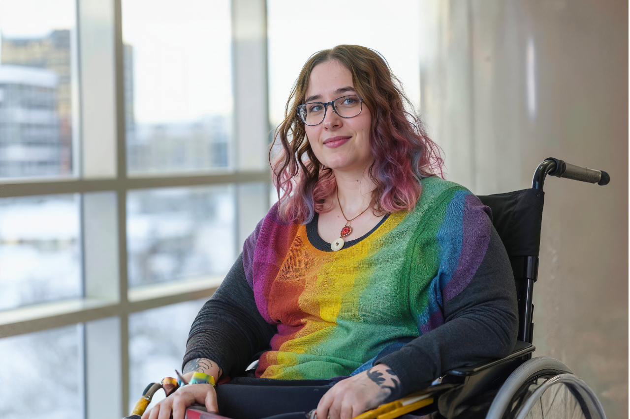 QueerPflege-LSBTIQ-Pflege Eine Person mit langen, welligen Haaren und Brille sitzt in einem Rollstuhl und trägt ein regenbogenfarbenes Oberteil. Im Hintergrund sind große Fenster mit einer städtischen Landschaft davor zu sehen, was die Bedeutung von queersensibler Pflege für die Schaffung integrativer Umgebungen unterstreicht.