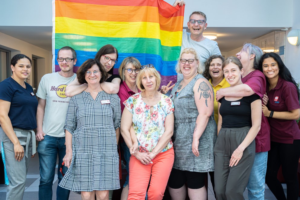 QueerPflege-LSBTIQ-Pflege Eine Gruppe von Menschen steht drinnen zusammen und hält eine Regenbogenflagge hoch. Sie lächeln und scheinen Teil einer unterstützenden und integrativen Gemeinschaft zu sein.