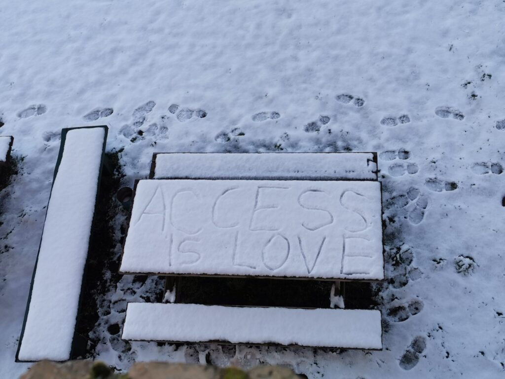 QueerPflege-LSBTIQ-Pflege Eine schneebedeckte Picknickbank mit den Worten „ACCESS IS LOVE“ im Schnee, umgeben von Fußabdrücken.