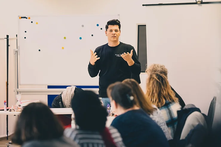 QueerPflege-LSBTIQ-Pflege Eine Person spricht vor einer Gruppe in einem Klassenzimmer. Hinter ihr steht eine Tafel. Mehrere Personen sitzen und blicken auf den Sprecher.