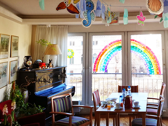 QueerPflege-LSBTIQ-Pflege Ein hell dekorierter Raum mit einem Klavier, einem Tisch und Stühlen. Im Fenster sind ein großer Regenbogen und verschiedene hängende Papierbasteleien zu sehen. Der Tisch ist mit Tassen, einem Topf und Wasserflaschen gedeckt.