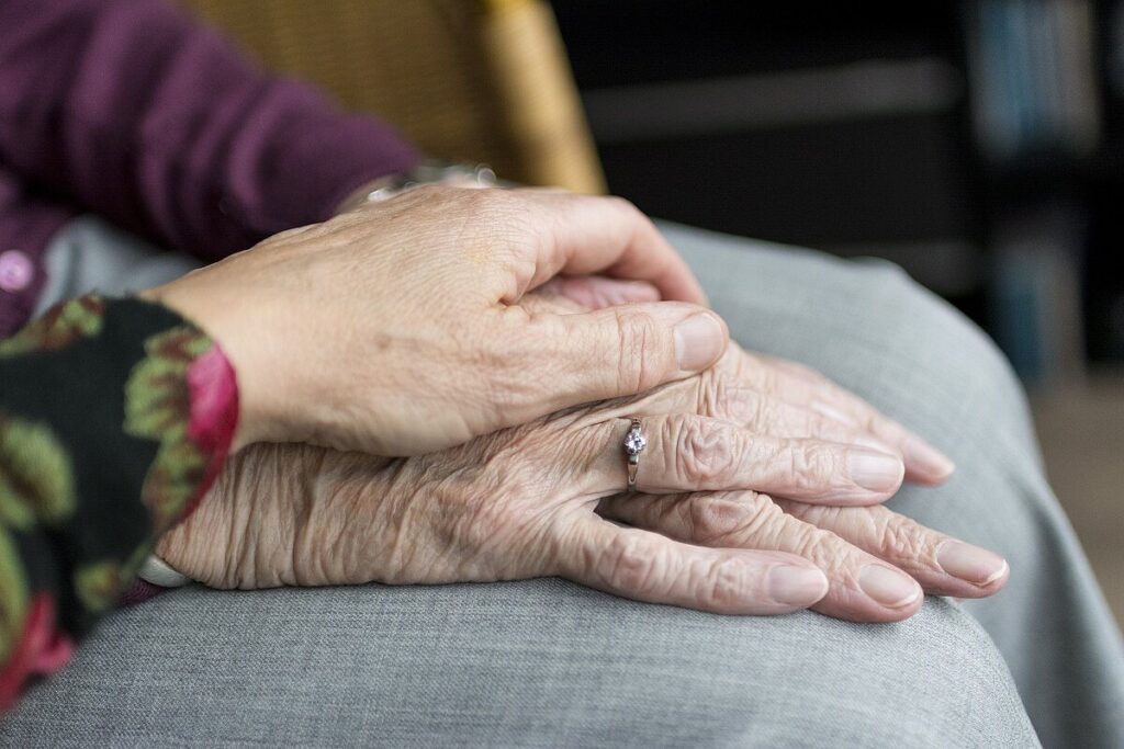 QueerPflege-LSBTIQ-Pflege Eine jüngere Hand ruht sanft auf einer älteren Hand mit einem Diamantring, beide Hände ruhen auf einer grauen Stoffoberfläche.