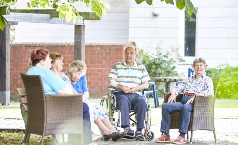 QueerPflege-LSBTIQ-Pflege Eine Gruppe älterer Menschen, einige sitzen auf Stühlen und eine Person im Rollstuhl, entspannt im Freien unter einem Baum.