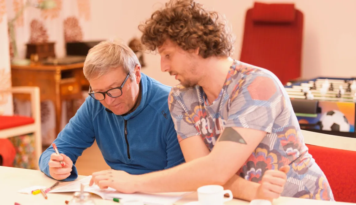 QueerPflege-LSBTIQ-Pflege Zwei Männer sitzen an einem Tisch und diskutieren oder lernen. Ein Mann mit Brille schreibt, während der andere mit lockigem Haar auf ein Dokument zeigt. Im Hintergrund sind verschiedene Gegenstände und ein Tischfußballtisch zu sehen.