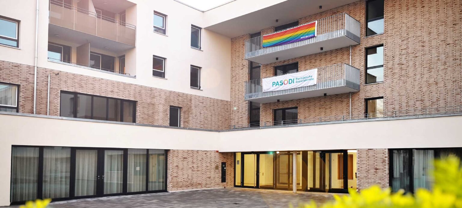 QueerPflege-LSBTIQ-Pflege Ein modernes Apartmentgebäude mit mehreren Balkonen, an denen eine Regenbogenflagge und ein Banner mit der Aufschrift „PASODI, Pastoral diocesana delle diversità sessuali“ hängen.