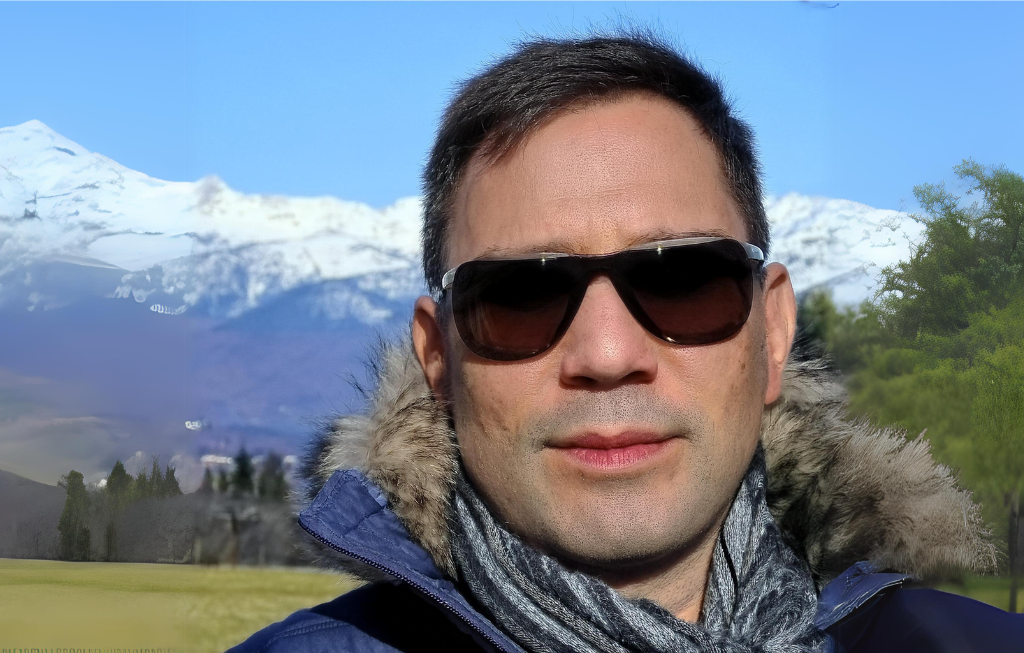 QueerPflege-LSBTIQ-Pflege Ein Mann mit Sonnenbrille und Schal steht im Freien mit schneebedeckten Bergen im Hintergrund.