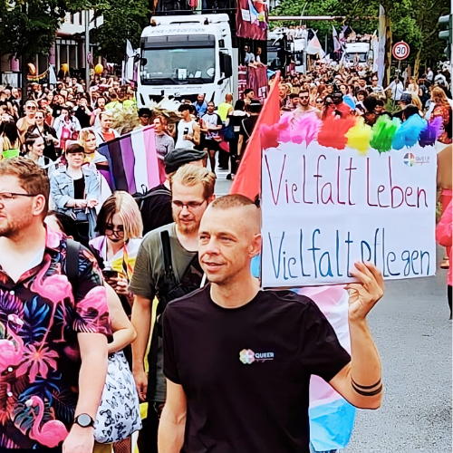 QueerPflege-LSBTIQ-Pflege Eine Menschenmenge bei einer Parade mit einer Person, die ein Schild mit der Aufschrift „Vielfalt leben, Vielfalt pflegen“ hochhält.